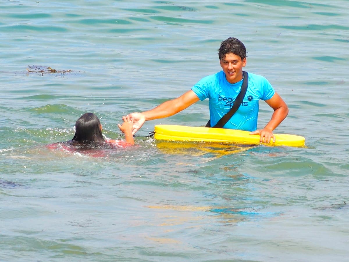 A BeachSports coach on a board staying right beside a young camper swimming in the calm ocean.