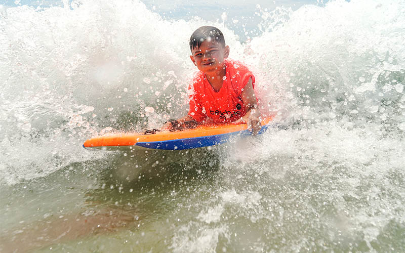 A young boy wearing an orange Beach Sports rash guard rides the white water on a boogie board at a kids summer beach camp in redondo beach
