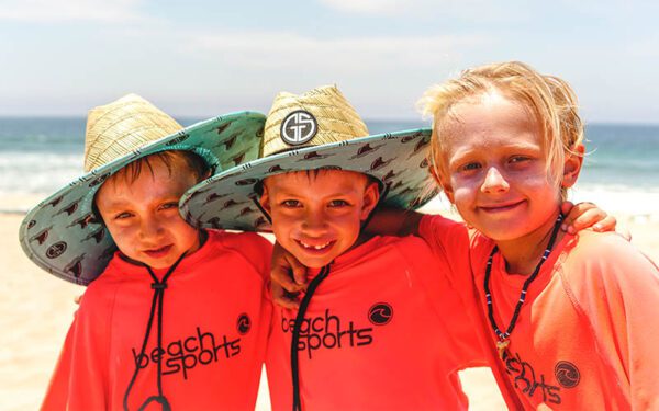 Three boys arm in arm at Hermosa Beach Sports summer camp smiling at the beach in the sun. Wearing sunscreen, wide brimmed hats and protective rash guards