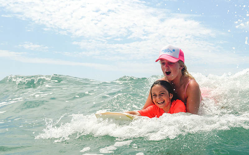 A beach sports kids camp instructor and camper have fun riding a wave together.